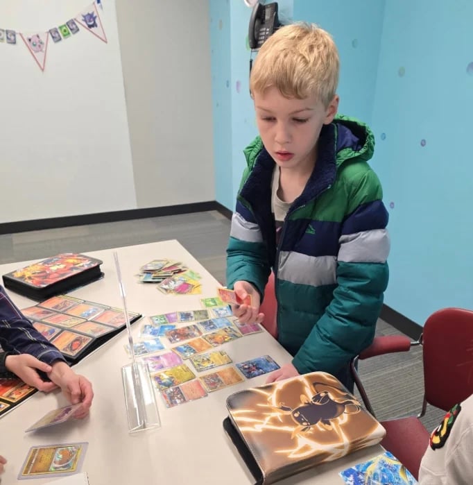 Young child with blonde hair wearing a green and navy jacket examining trading cards at a table indoors