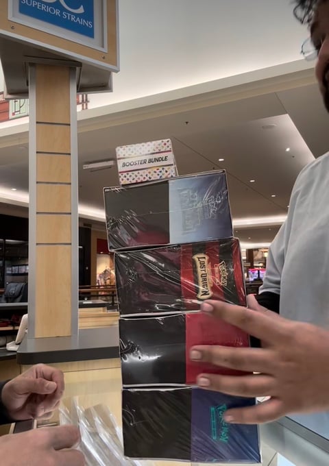 Person holding stacked video game cases labeled Booster Bundle in a shopping mall, with store displays visible in background