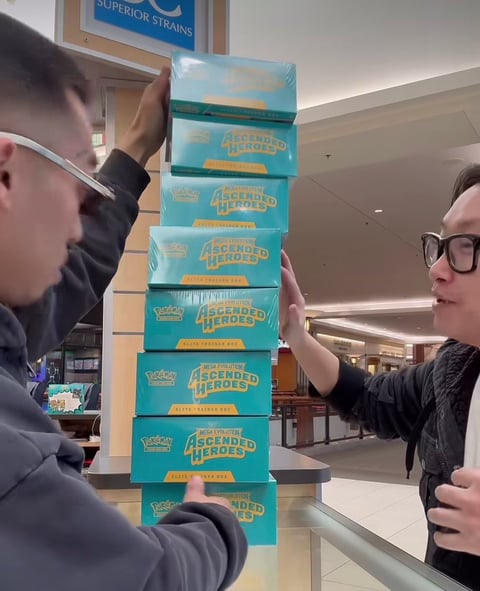 Two people stacking turquoise trading card game booster boxes in a precarious tower indoors